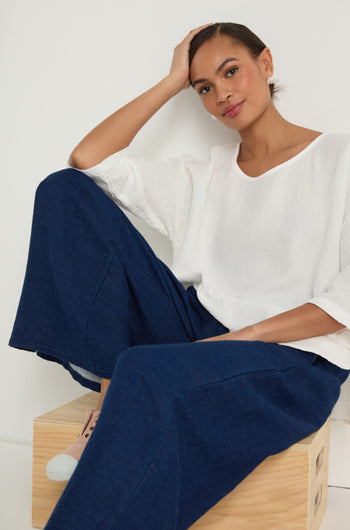 A woman wearing Wide Leg Slit Trousers and a white blouse sits on a wooden box, resting one hand on her head and looking at the camera.
