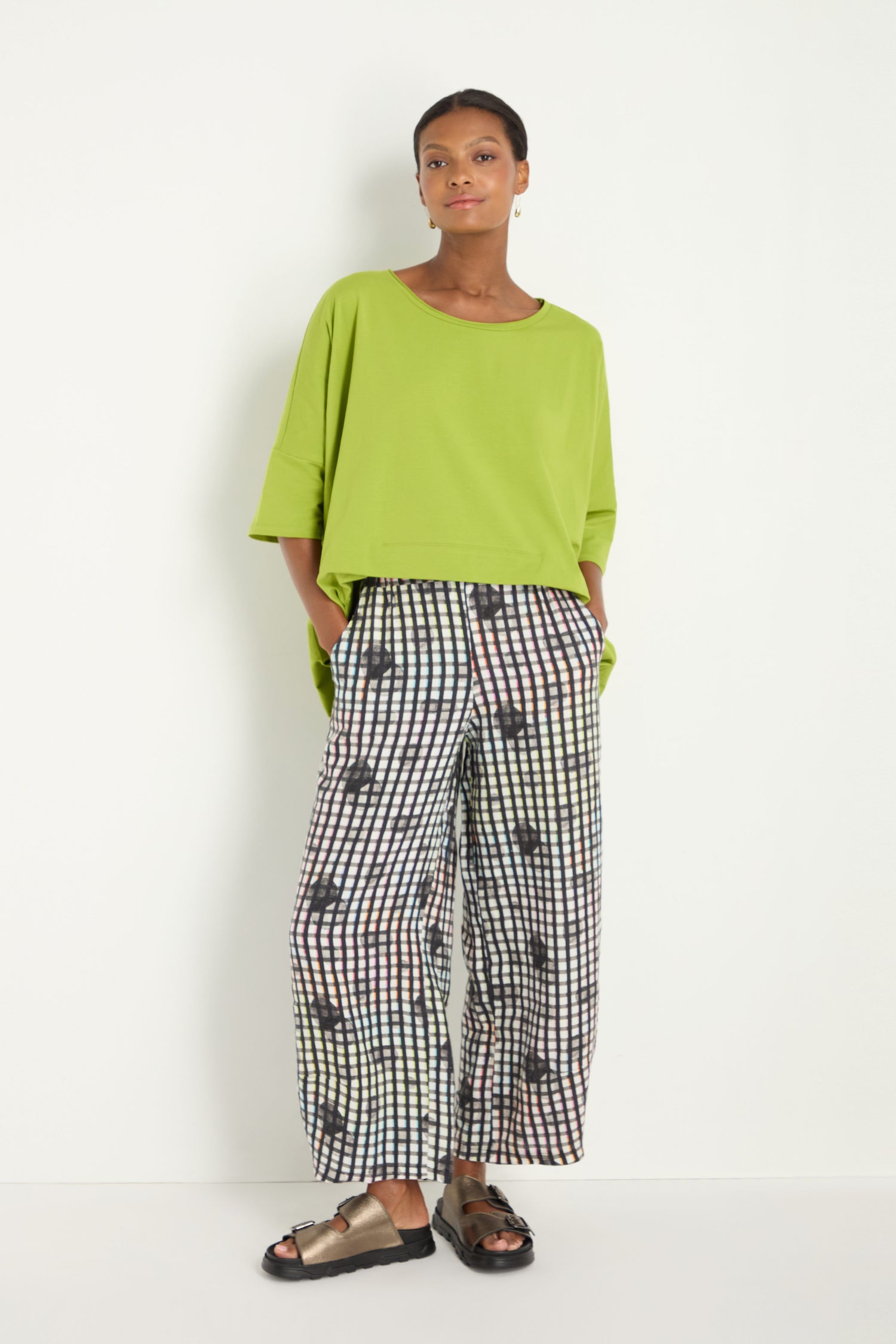 A woman stands against a white wall wearing a loose lime green top, Check Dot Linen Trousers with abstract patterns, and black sandals.
