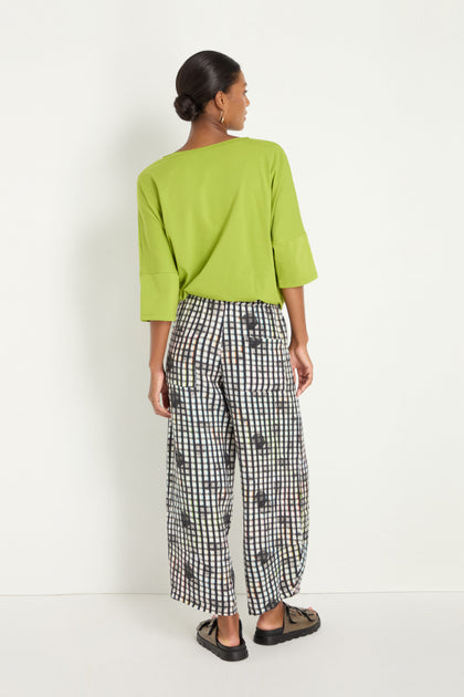 A woman faces away in a loose lime green top, Check Dot Linen Trousers with a relaxed straight-leg fit and grid check pattern, and black sandals against a plain white background.