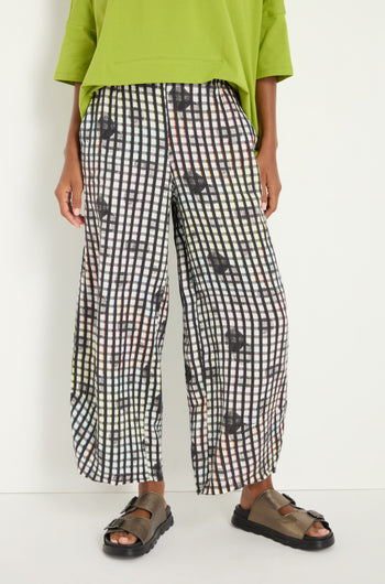 A person stands against a plain white background wearing a loose lime green top, brown sandals, and black and white Check Dot Linen Trousers with a relaxed straight leg.