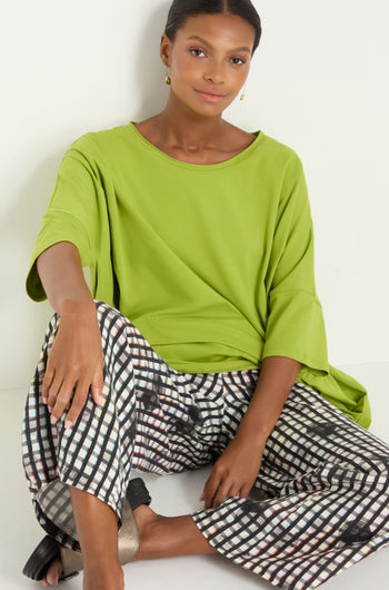 Woman sitting against a white wall, wearing a loose lime green top, Check Dot Linen Trousers, gold earrings, and silver sandals.