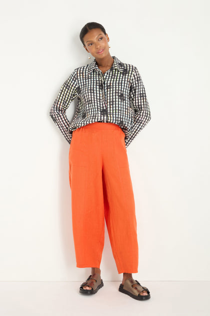 A person stands against a white wall wearing a black-and-white plaid shirt, Linen Bubble Trousers in burnt orange, and black sandals.