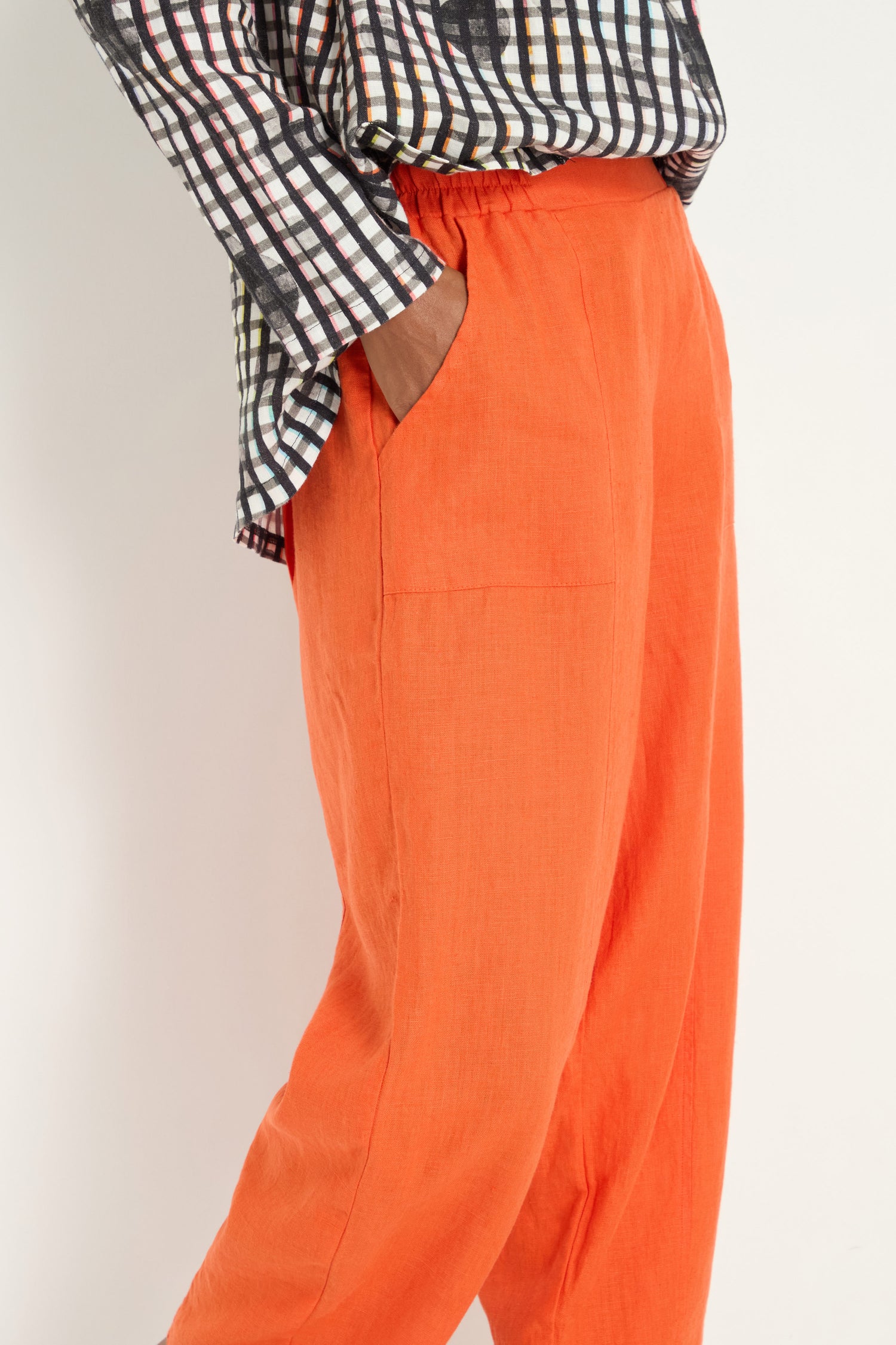 Someone wears Linen Bubble Trousers in burnt orange with side pockets and a black-and-white checkered long-sleeve shirt, standing with one hand in their pocket.