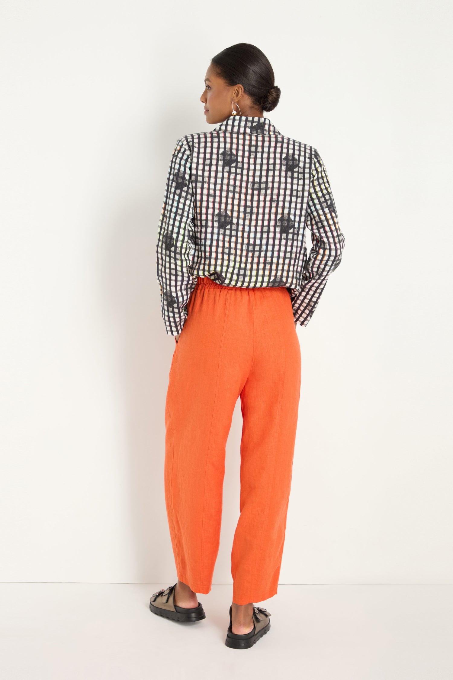 A person stands facing away, wearing an untucked black-and-white checkered shirt, Linen Bubble Trousers in burnt orange, and black shoes, set against a plain white background.