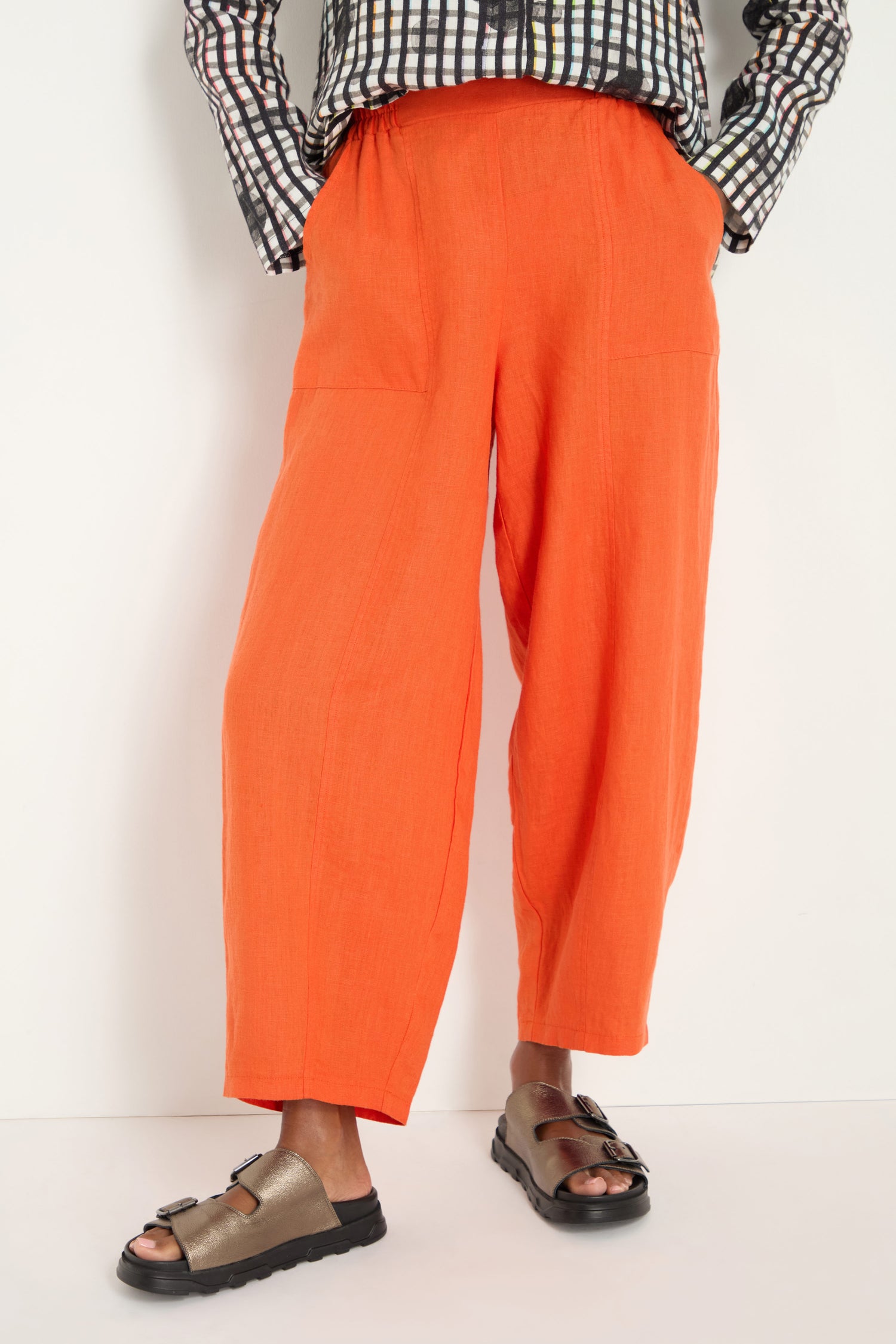 A person stands against a white background, wearing bright burnt orange Linen Bubble Trousers, a black-and-white patterned shirt, and metallic sandals.