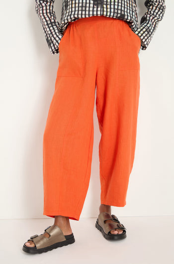 A person stands against a white background, wearing bright burnt orange Linen Bubble Trousers, a black-and-white patterned shirt, and metallic sandals.