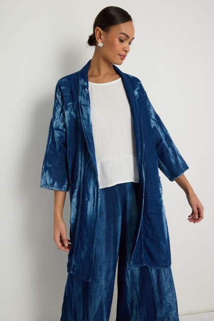 A woman stands against a plain white wall, wearing the Soft Denim Easy Jacket in a relaxed blue tie-dye style over a white top, paired with matching pants.