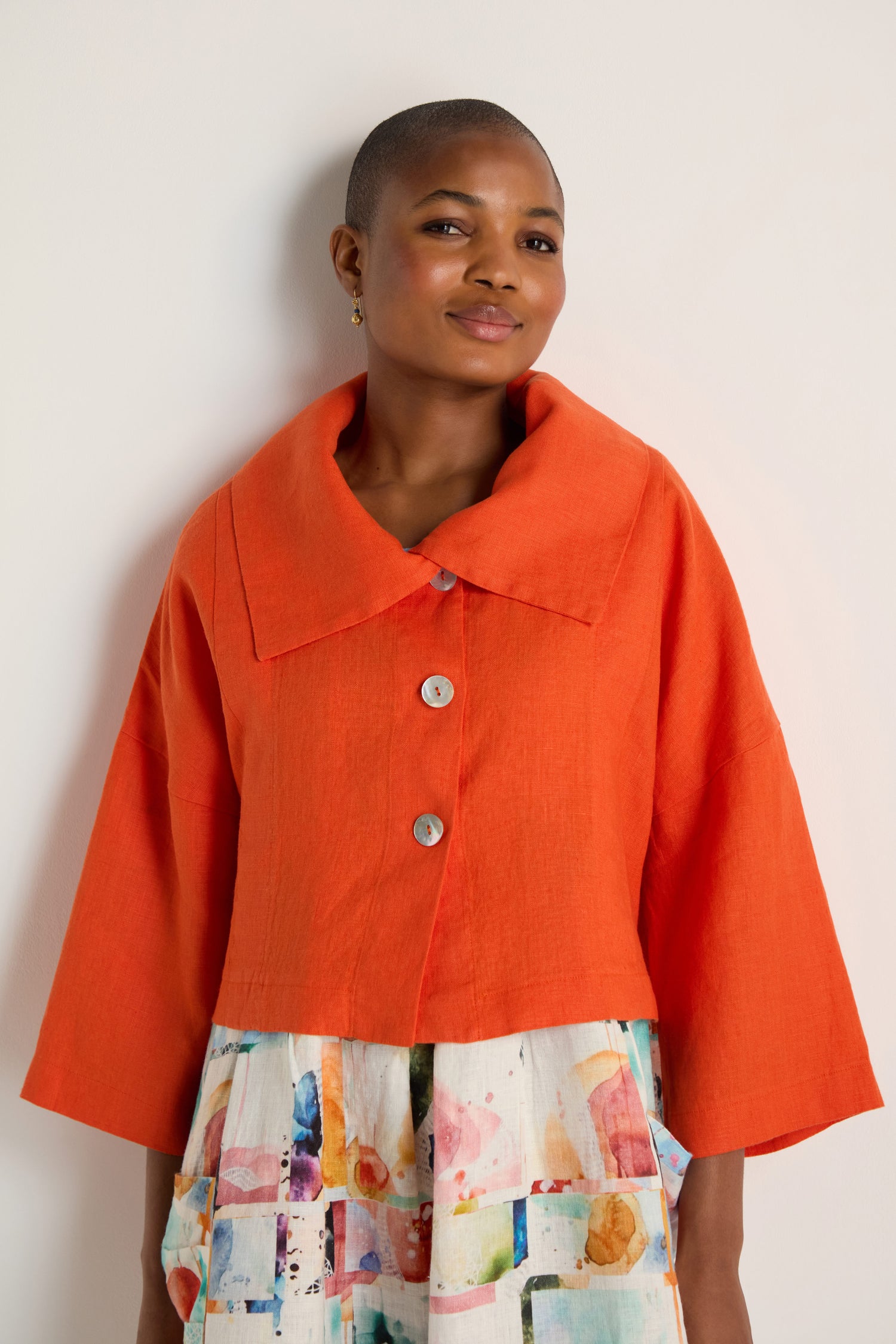 A woman stands against a plain light background, wearing the Wide Collar Linen Jacket in burnt orange—a cropped, boxy style—over a white dress with a colorful abstract print.