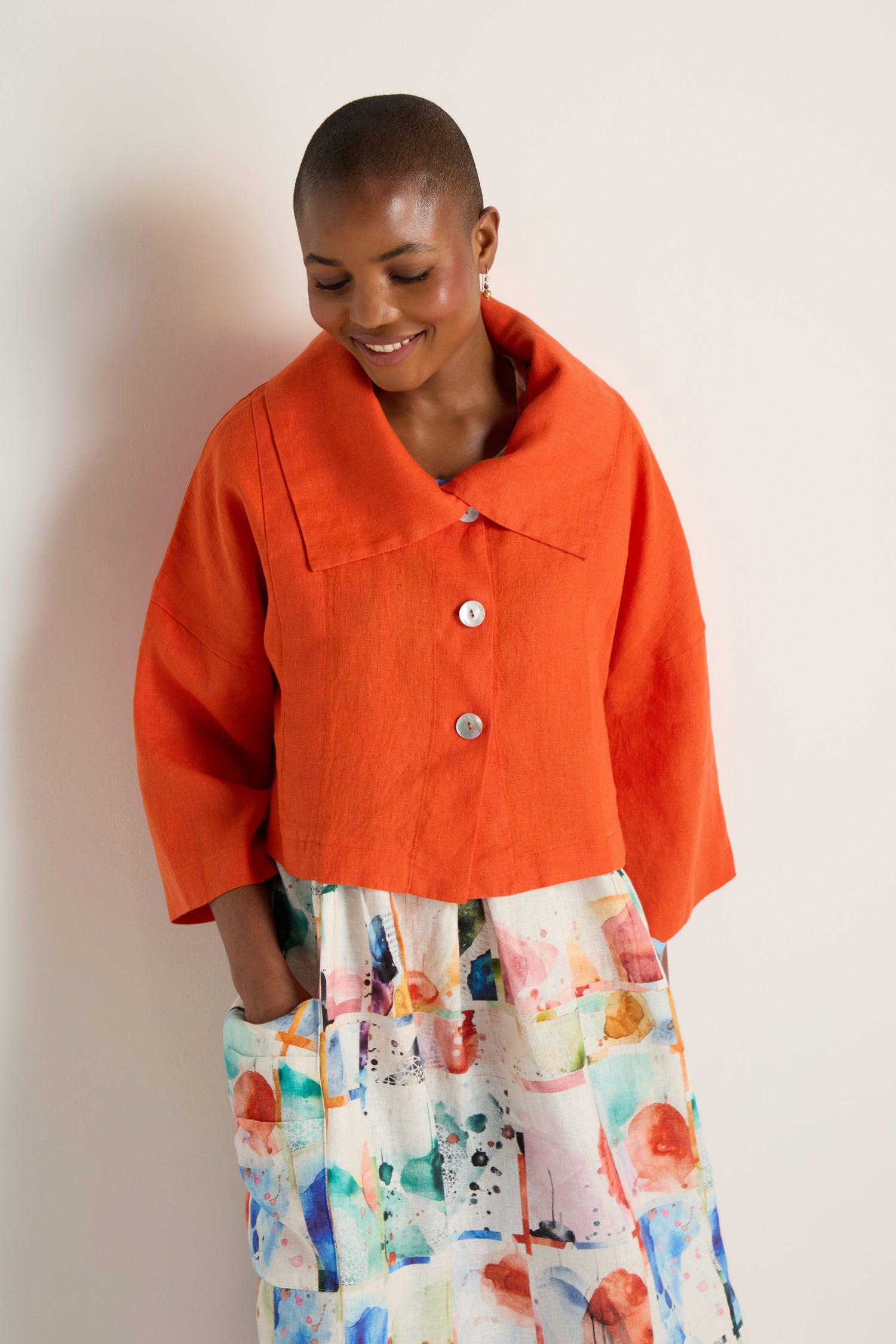 A person wears a Wide Collar Linen Jacket over a colorful, abstract-patterned dress, standing against a plain light background and smiling with hands in the dress pockets.