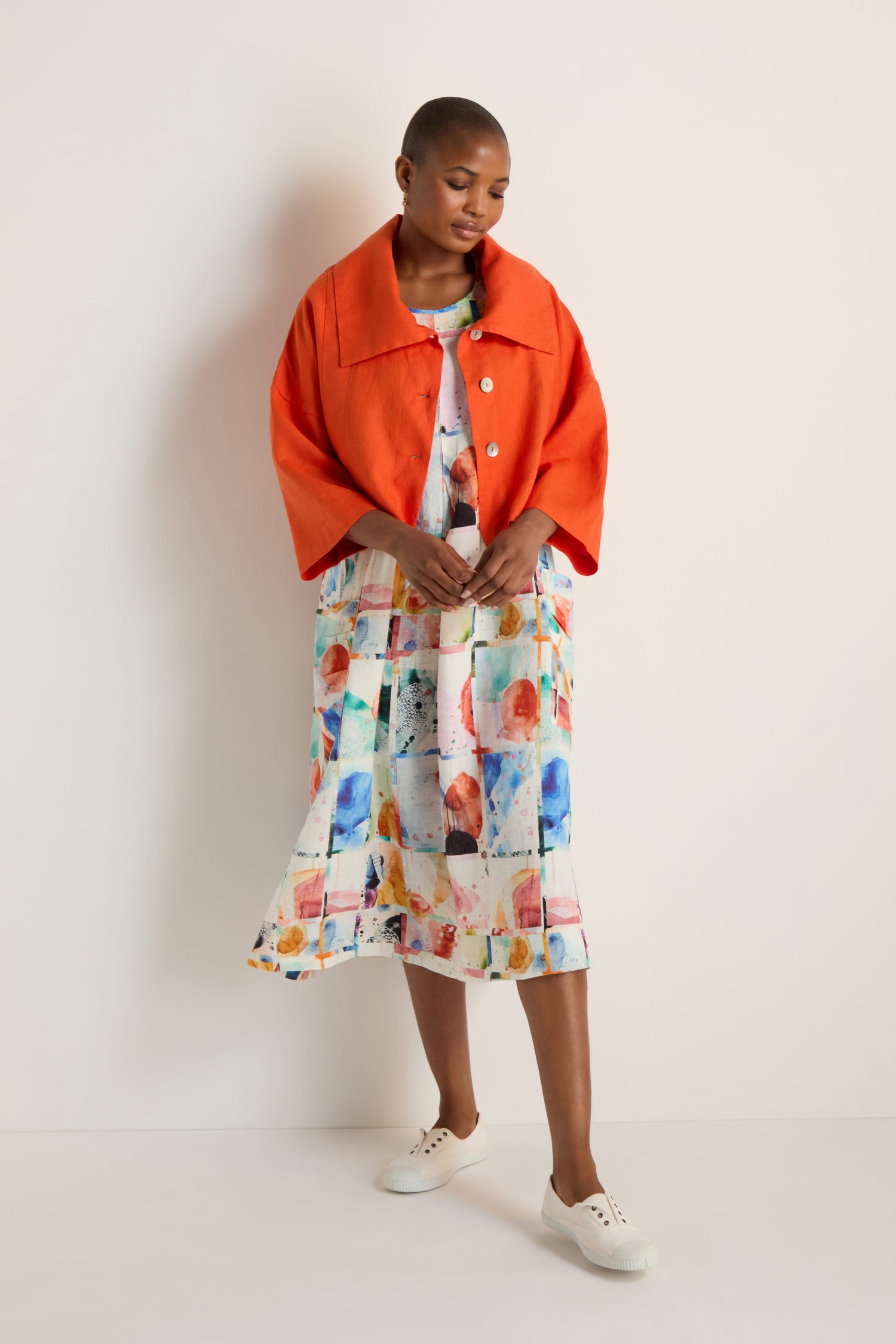 A person wearing a bright orange Wide Collar Linen Jacket over a colorful, abstract-patterned dress and white sneakers stands against a plain light background.