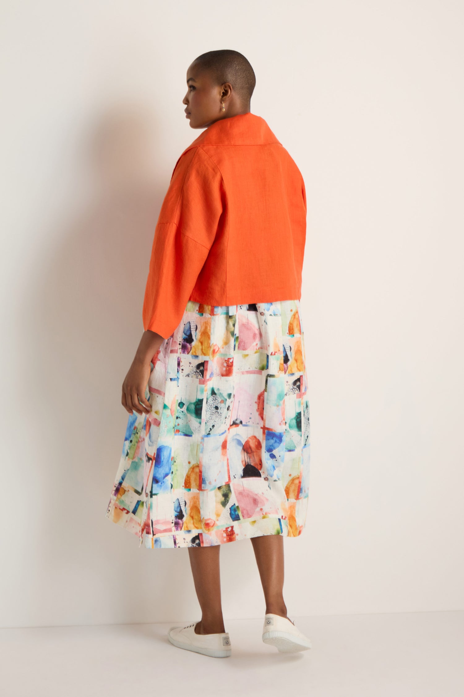 A person stands against a light wall, facing slightly away from the camera, wearing a bright orange Wide Collar Linen Jacket, a colorful abstract-print skirt, and white sneakers.