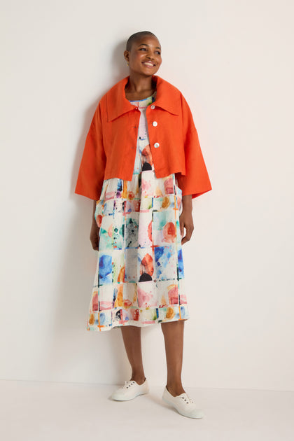 A person stands against a plain wall, wearing a burnt orange Wide Collar Linen Jacket over a colorful abstract midi dress and white sneakers.