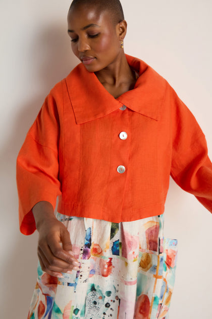 A person wearing a Wide Collar Linen Jacket in bright orange and a white skirt with a colorful abstract pattern stands against a plain background.