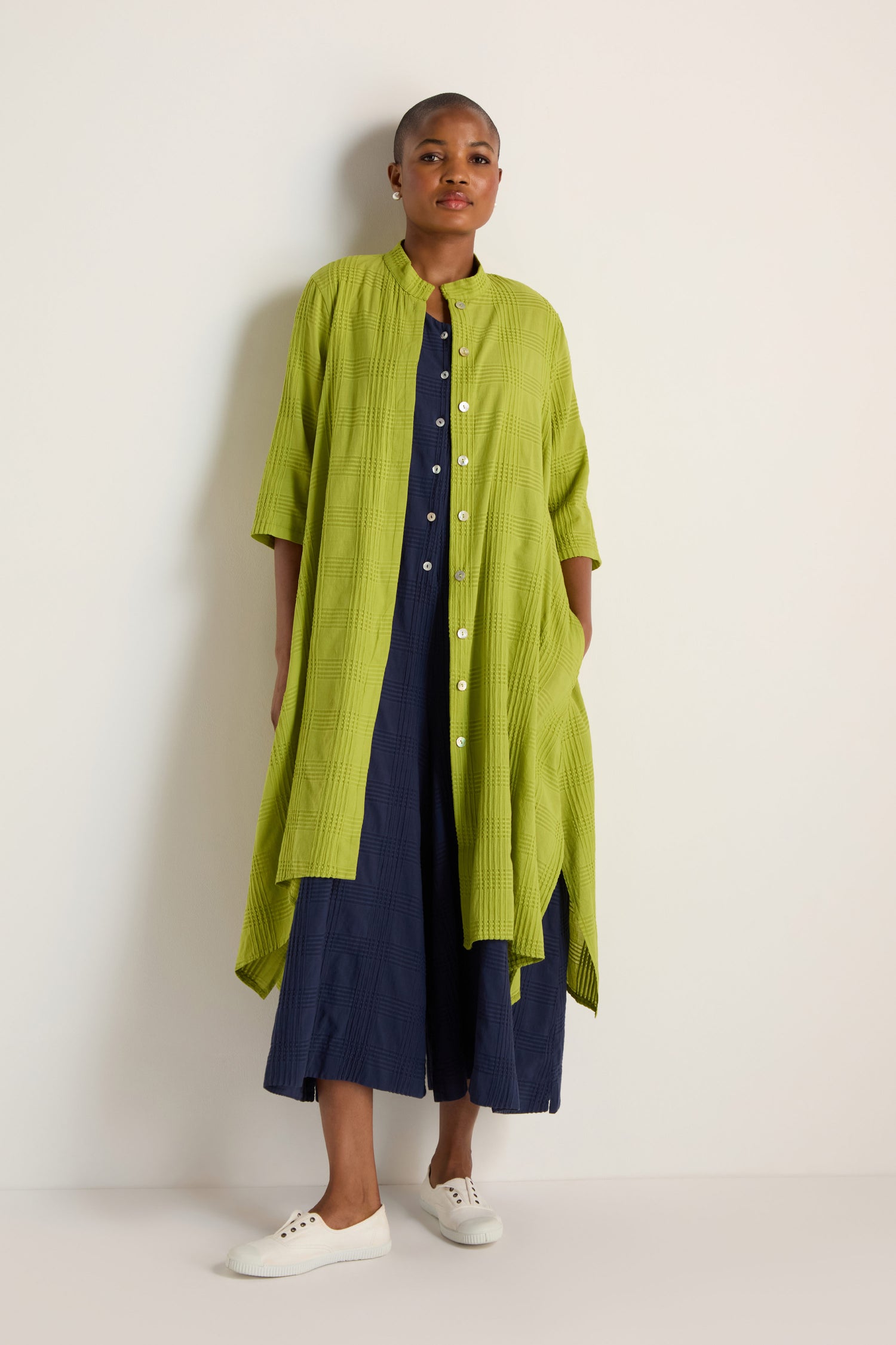 A person stands against a white wall wearing the Self Check Lime Shirt Dress over a navy dress and white sneakers.