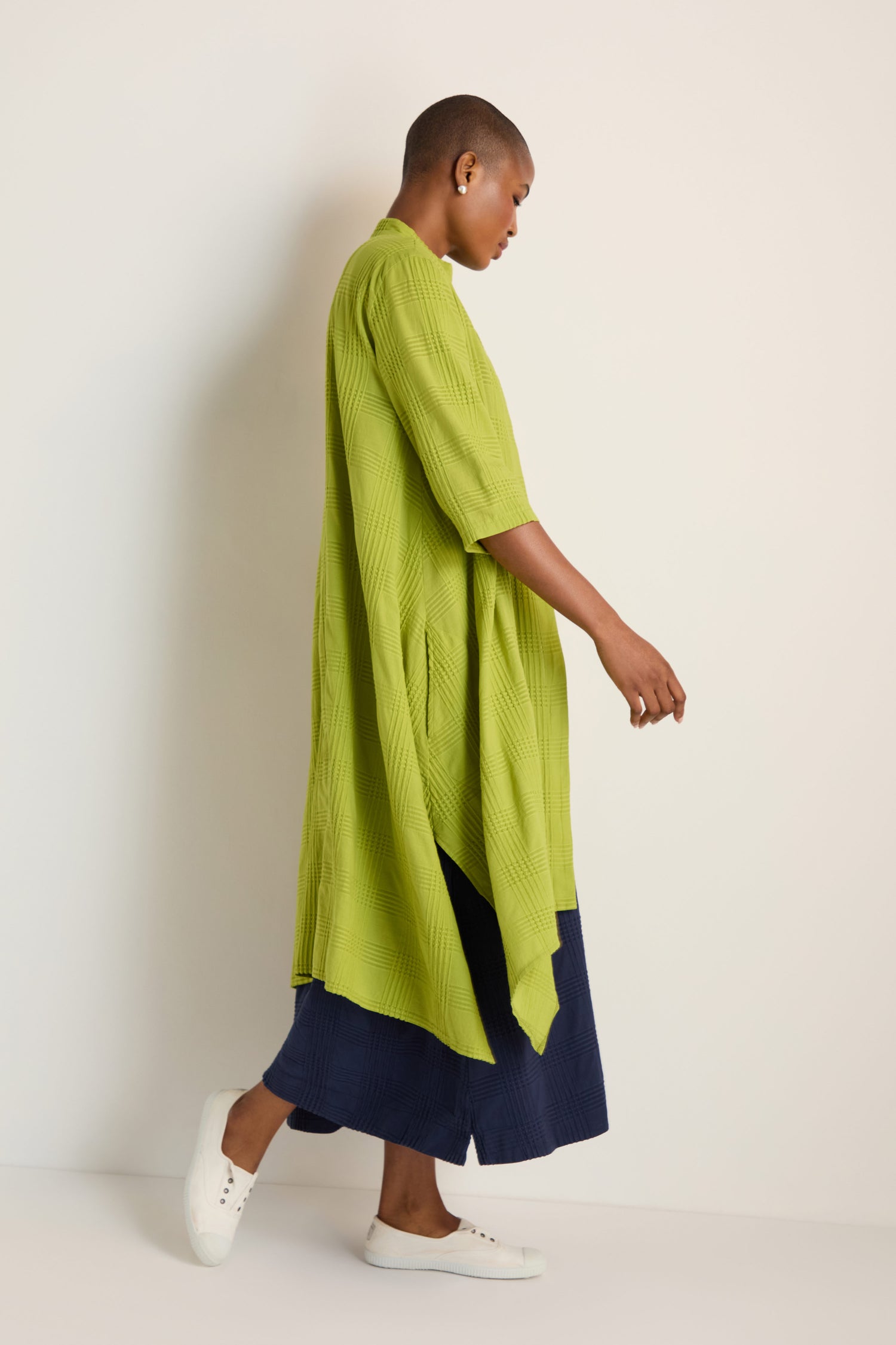 A person stands sideways in the Self Check Lime Shirt Dress styled as a long flowy top, paired with navy wide-leg pants and white sneakers against a plain background.