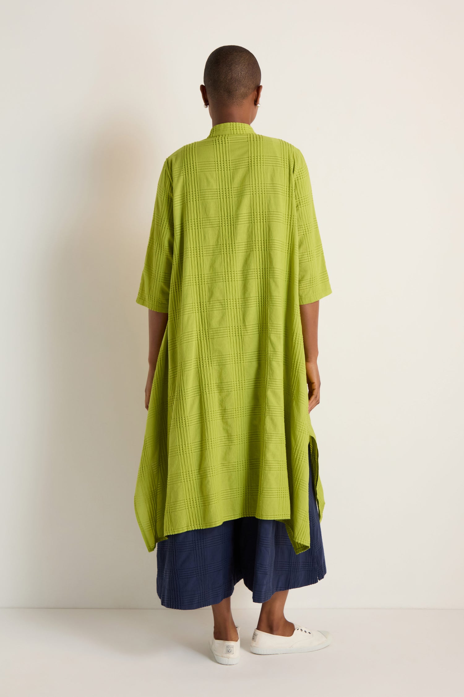 A person is seen from behind against a light background, wearing the Self Check Lime Shirt Dress, navy wide-leg pants, and white slip-on shoes.