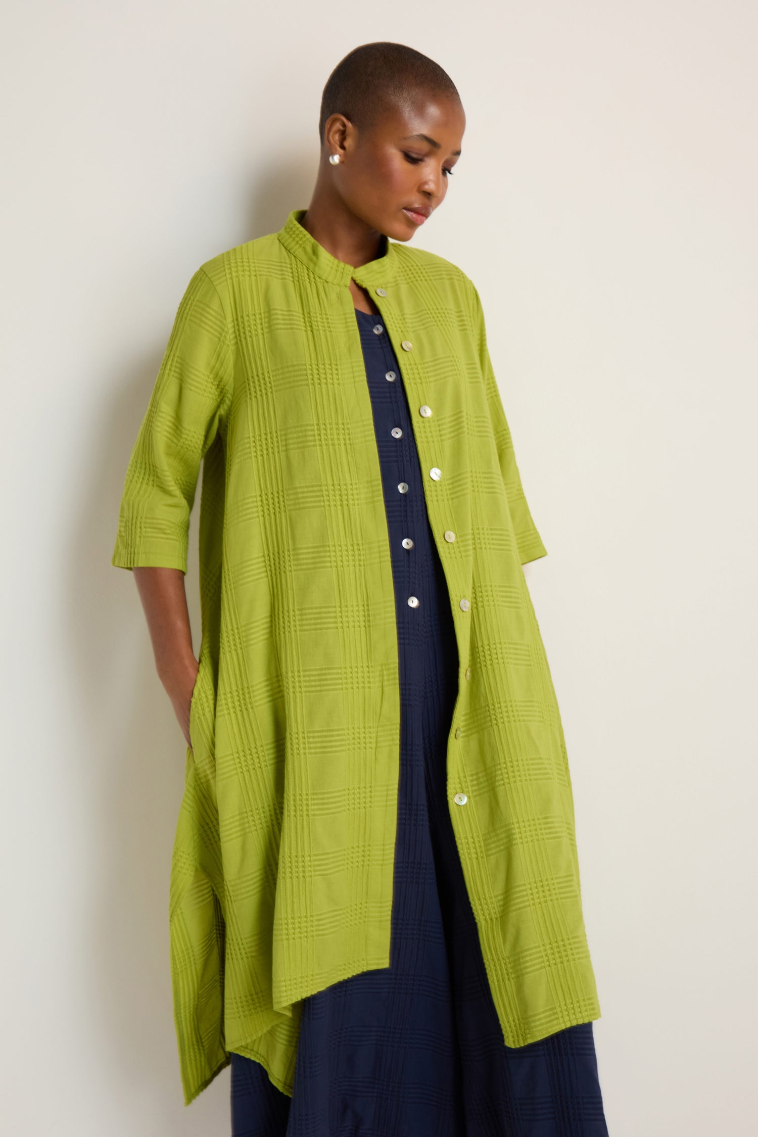 A person wears the Self Check Lime Shirt Dress with three-quarter sleeves, buttoned over a navy dress, standing against a plain light background.