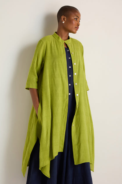 A person wearing the Self Check Lime Shirt Dress with elbow-length sleeves over a dark blue dress stands against a light-colored wall.