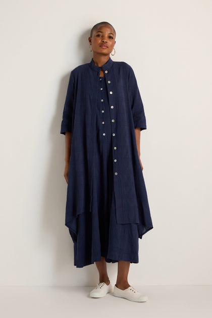 A person stands against a plain wall wearing the Self Check Navy Shirt Dress, which is loose-fitting with three-quarter sleeves, paired with white sneakers.