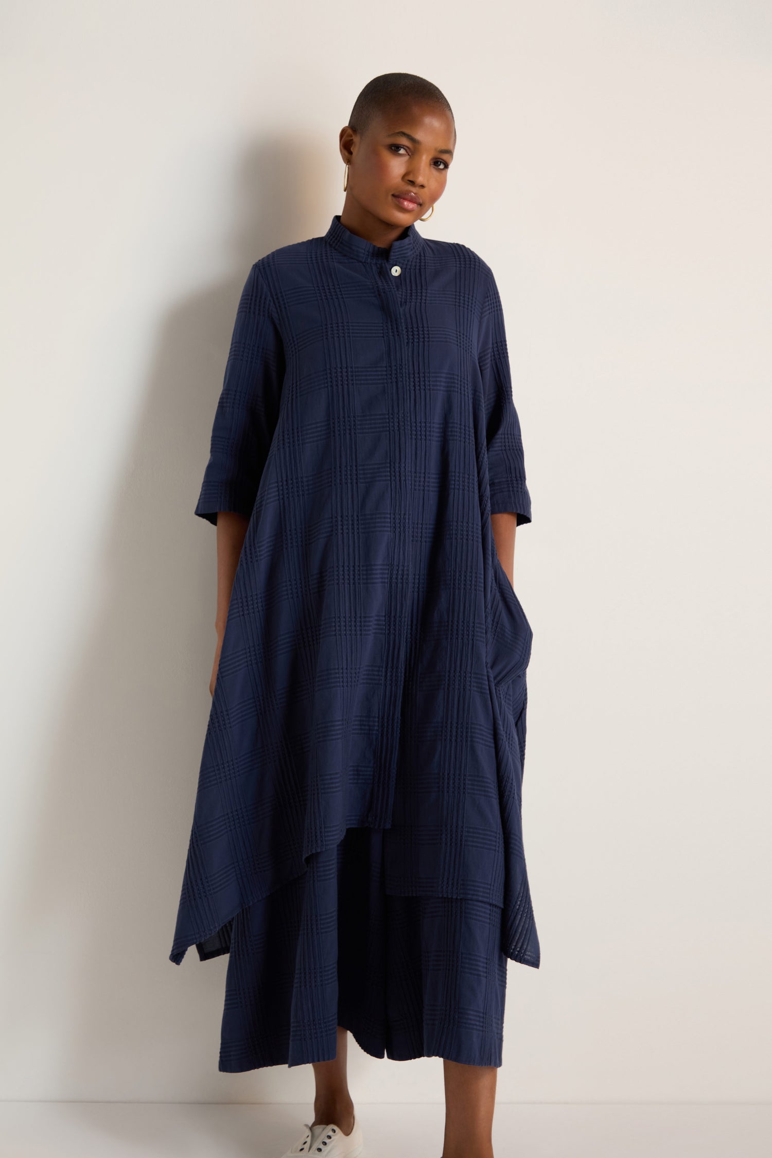 A person wearing the Self Check Navy Shirt Dress—loose-fit, long sleeves, high collar, and side pockets—stands against a plain light-colored wall.