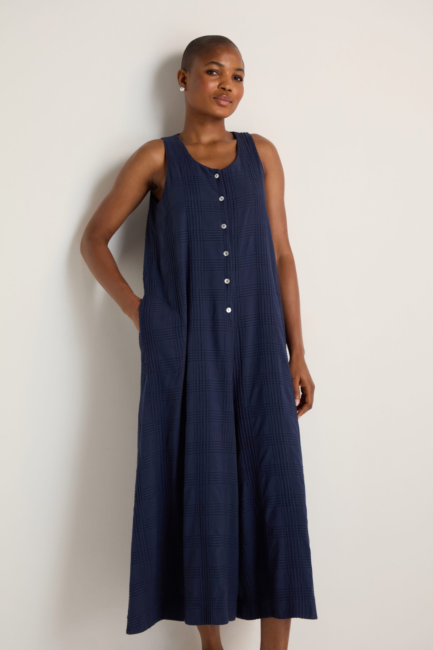 A person models the Self Check Sleeveless Jumpsuit, featuring a button-front design and navy textured fabric, standing by a plain light wall with one hand in the pocket.
