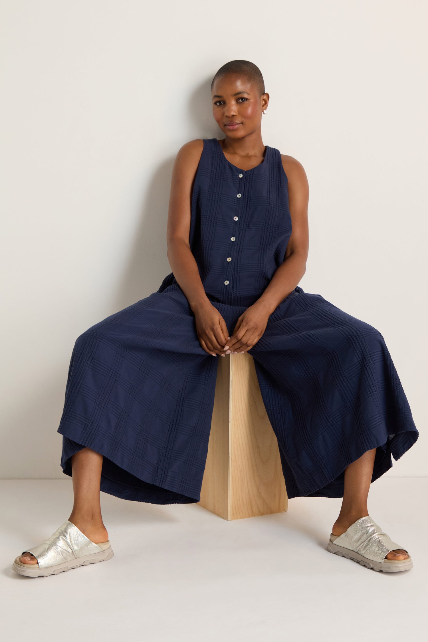 A person wearing the Self Check Sleeveless Jumpsuit sits on a wooden block against a plain white background.
