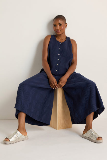 A person wearing the Self Check Sleeveless Jumpsuit sits on a wooden block against a plain white background.