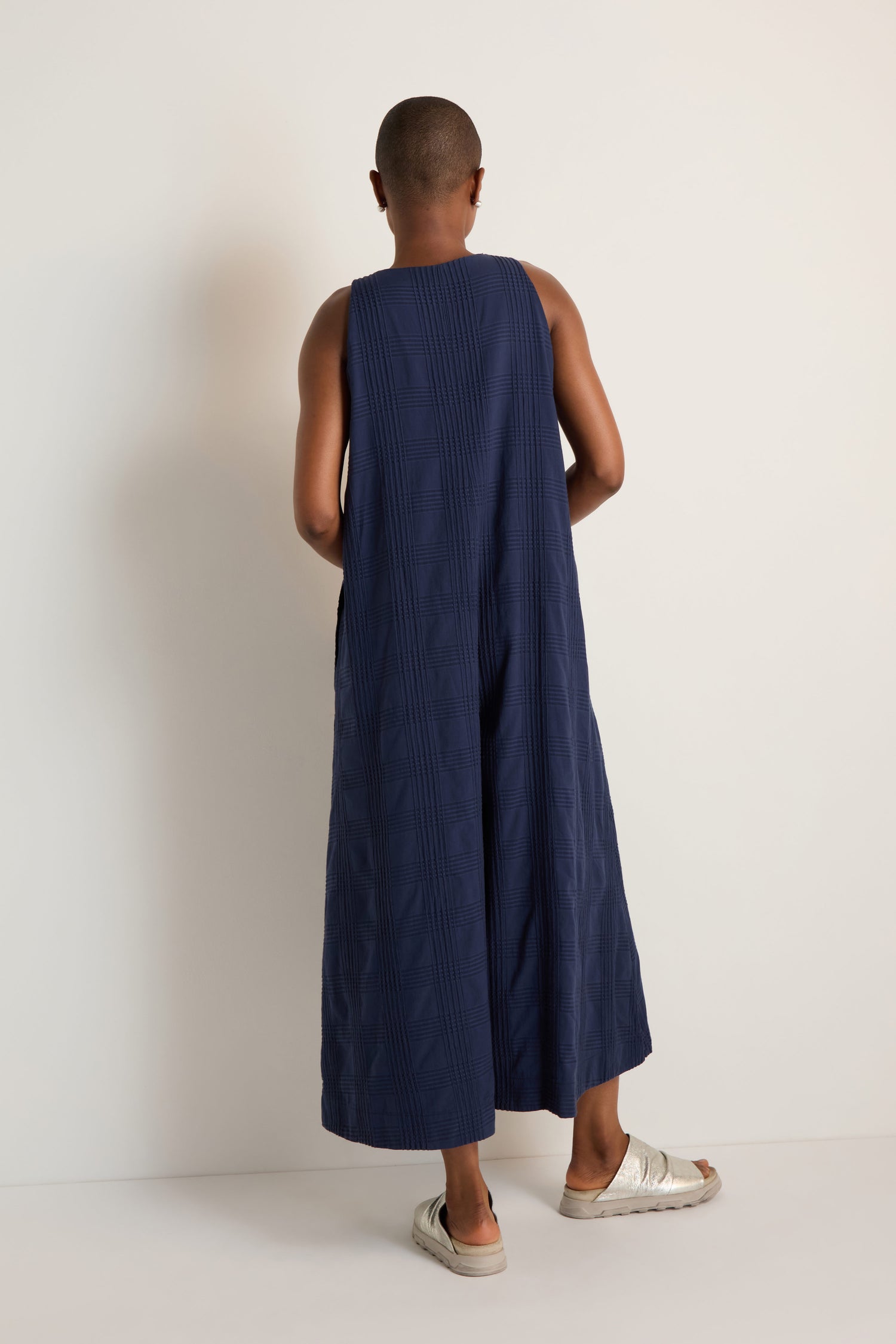 A person stands with their back to the camera, wearing the Self Check Sleeveless Jumpsuit in loose-fitting navy, paired with white sandals, against a plain light background.