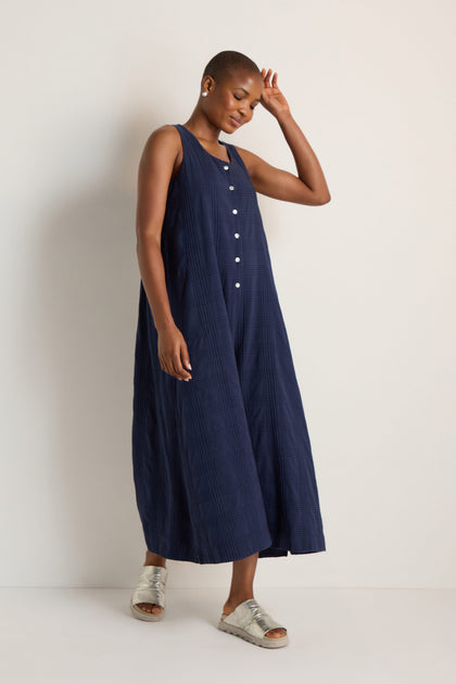 A person stands against a light background wearing the Self Check Sleeveless Jumpsuit, a loose-fitting navy cotton piece with front buttons, paired with silver sandals.