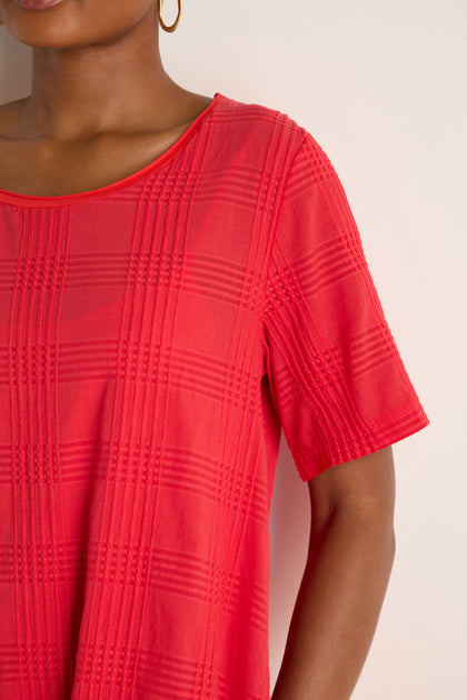 Close-up of a person wearing the Self Check Flared Dress in vibrant red textured cotton with short sleeves, a round neckline, gold hoop earrings, and a light background.