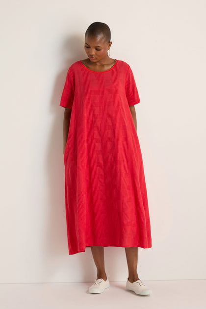 A person stands against a white wall wearing a loose-fitting, short-sleeved Self Check Flared Dress in red and white shoes.