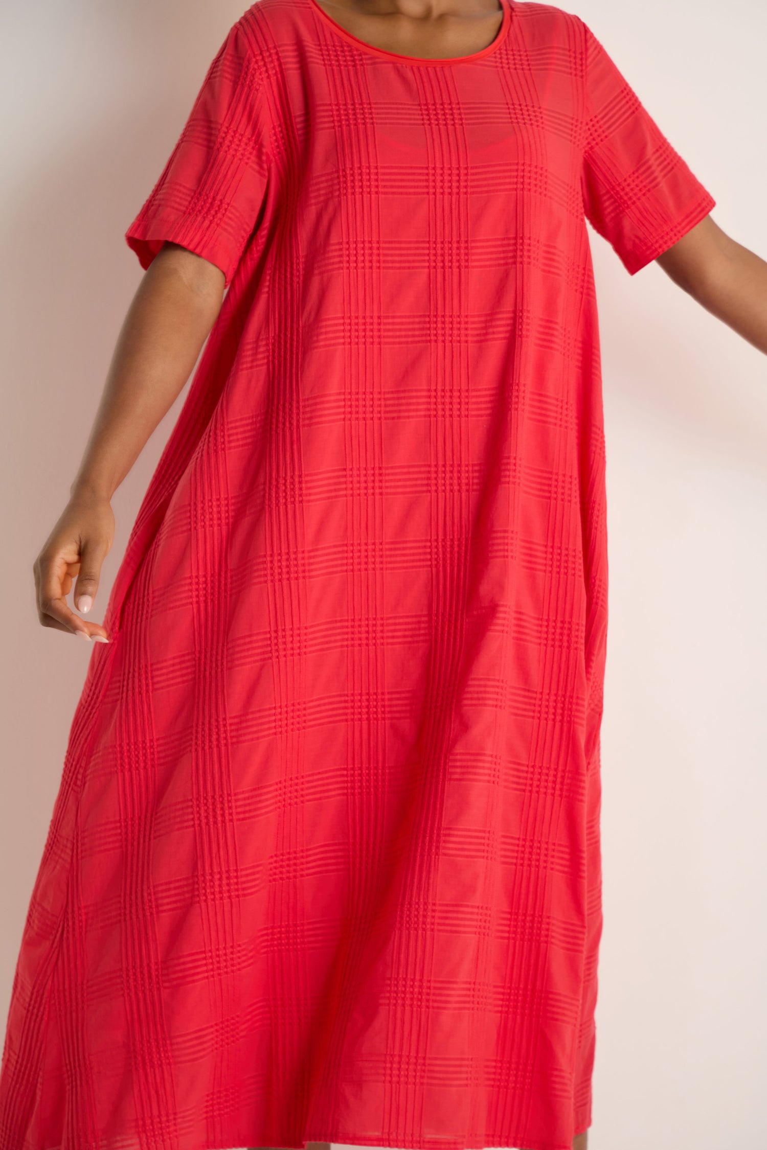 A person is shown from the torso up wearing a loose-fitting, short-sleeved Self Check Flared Dress in textured red cotton against a plain background.