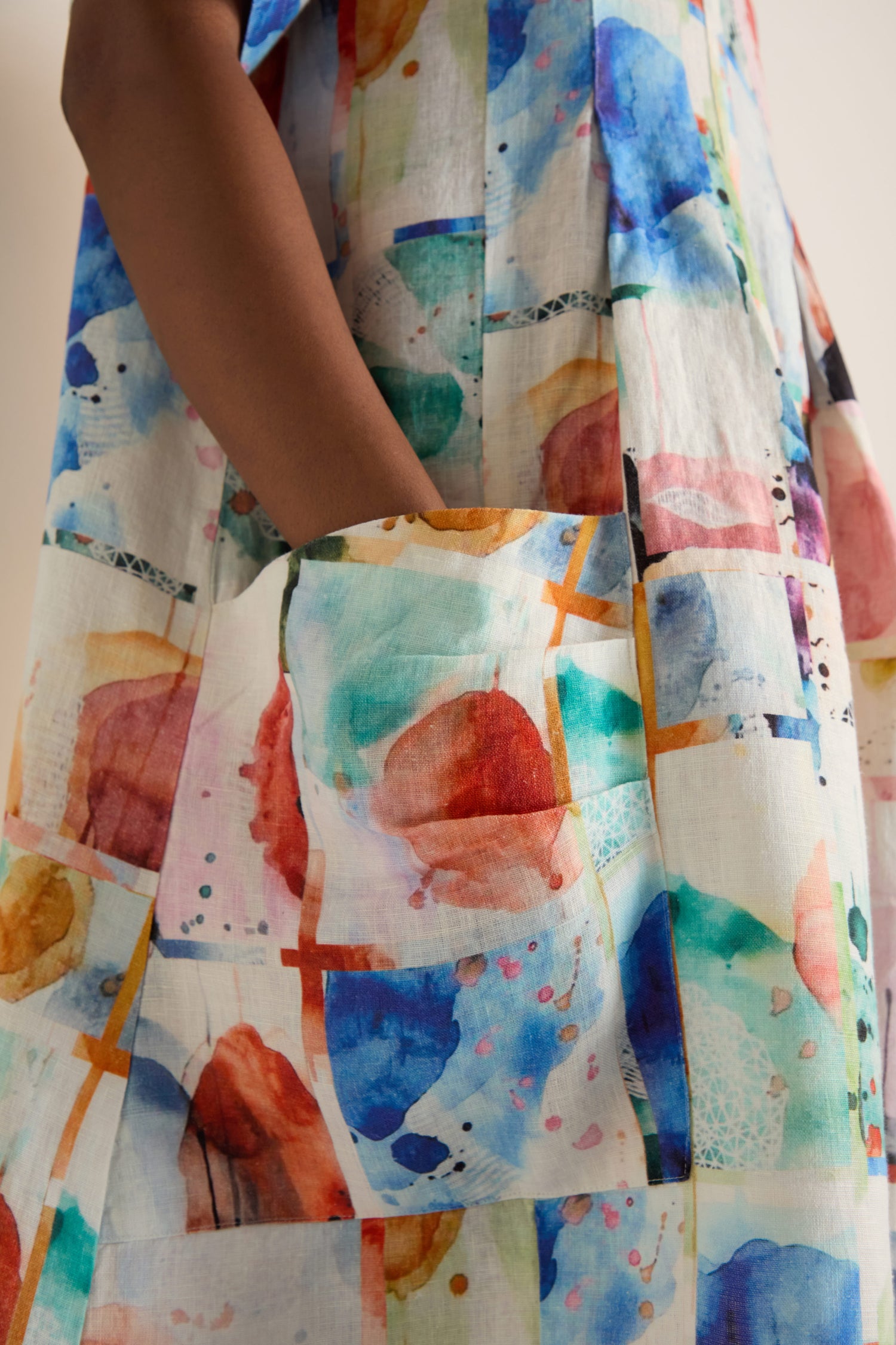 A person with medium brown skin wears the Watercolour Linen Pleated Dress, showcasing its colorful abstract pattern and places a hand inside a large pocket.