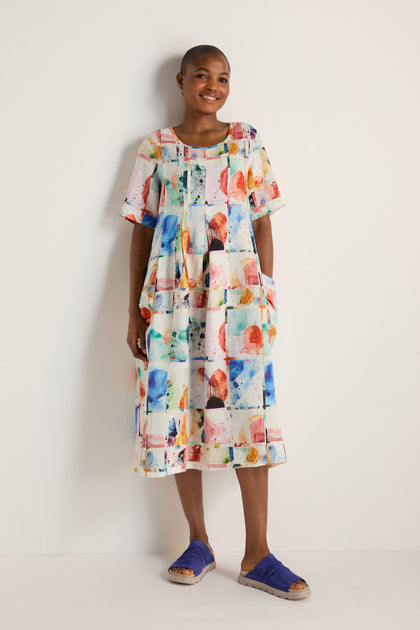 Standing against a white wall, the person smiles with hands in the pockets of the Watercolour Linen Pleated Dress—an airy, colorful summer dress with short sleeves—paired with blue slippers.