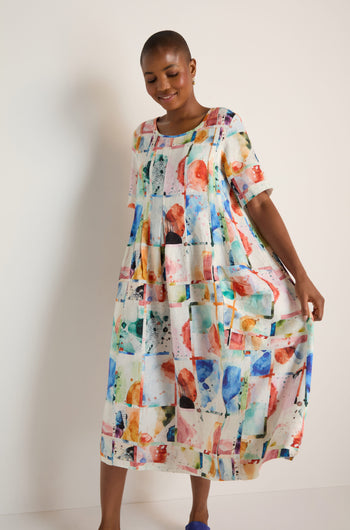 A person wears the Watercolour Linen Pleated Dress, featuring a vibrant abstract pattern, and stands against a white background smiling while holding out one side of the pleated midi dress.