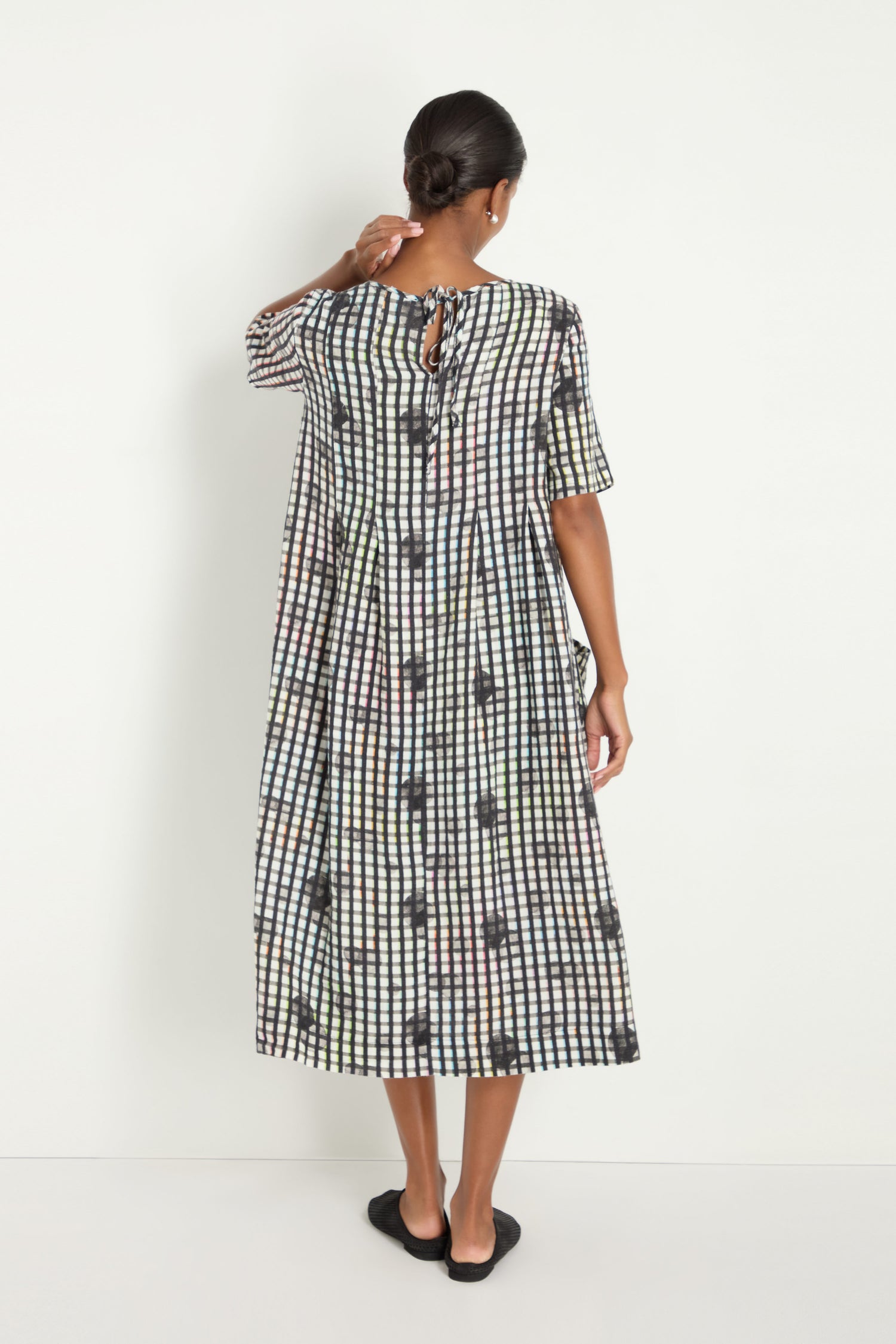 A woman with her hair in a bun stands facing a white wall, wearing the Check Dot Pleated Linen Dress with short sleeves and black flats.