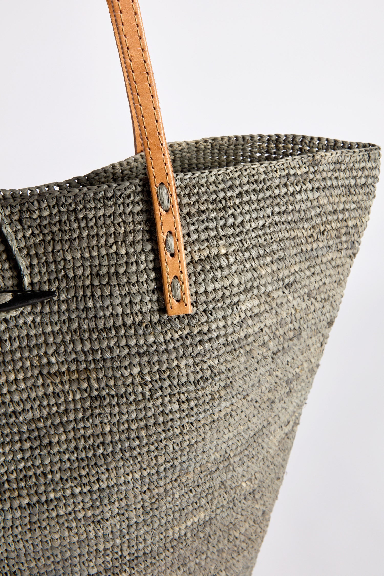 Close-up of the Ilana Raffia Basket, featuring its woven gray pattern and highlighted by a tan leather strap with metal rivets.