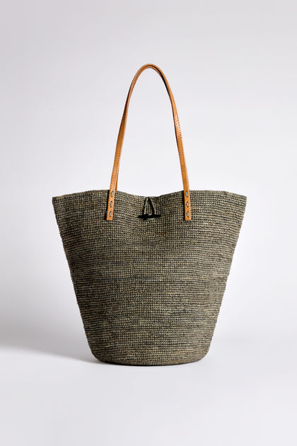 The Ilana Raffia Basket showcases handwoven raffia, complemented by rich brown leather handles, and a front decorative knot on a plain backdrop.