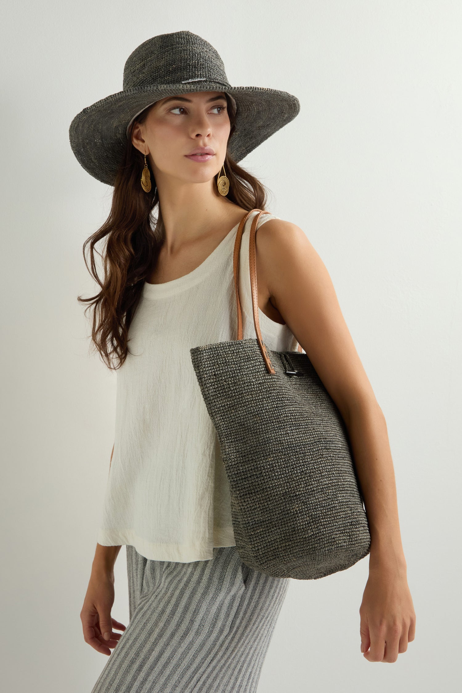 A woman in a white sleeveless top and striped pants, wearing a large gray hat, holds the Ilana Raffia Basket known for its spacious interior. Her long brown hair complements her drop earrings beautifully.