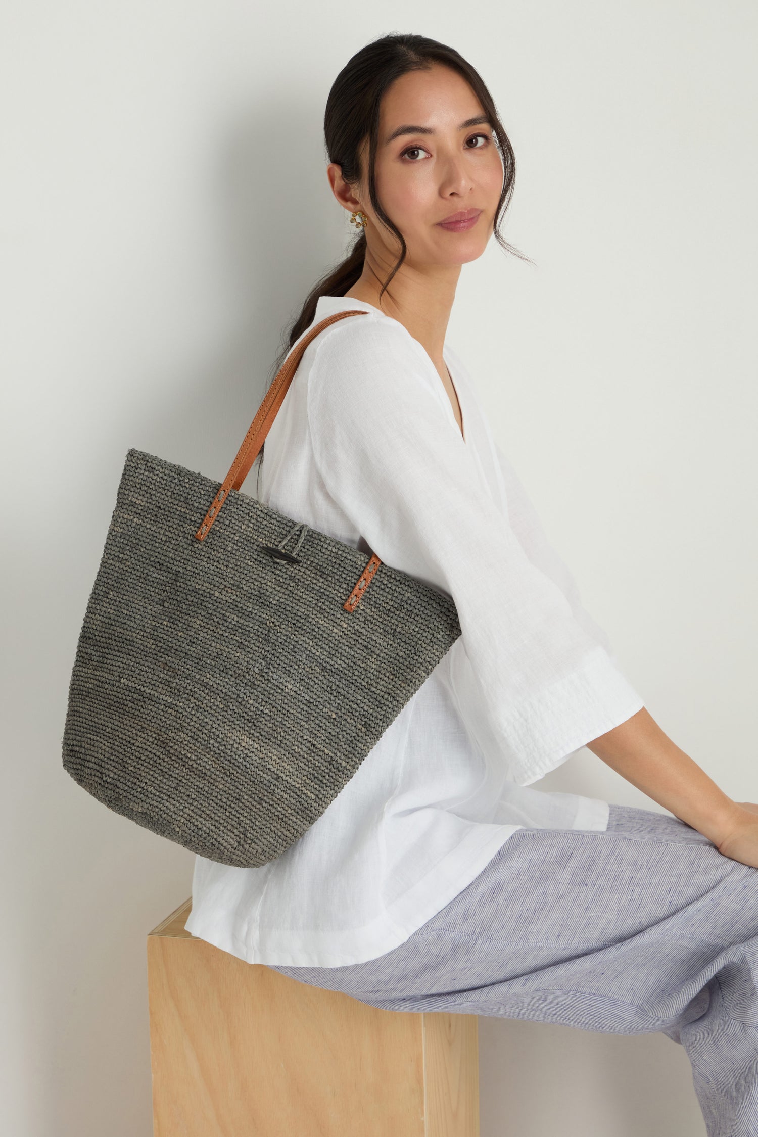 A woman sits on a wooden block, wearing a white blouse and light gray pants, carrying the Ilana Raffia Basket tote with brown handles over her shoulder.
