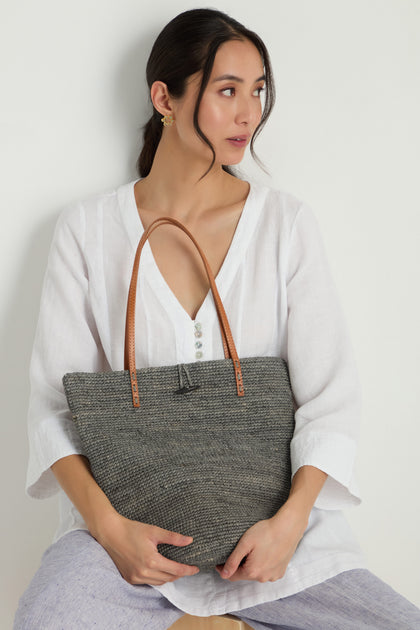 Woman in a white blouse sits against a white wall, holding the Ilana Raffia Basket large gray woven tote with brown handles on her lap.