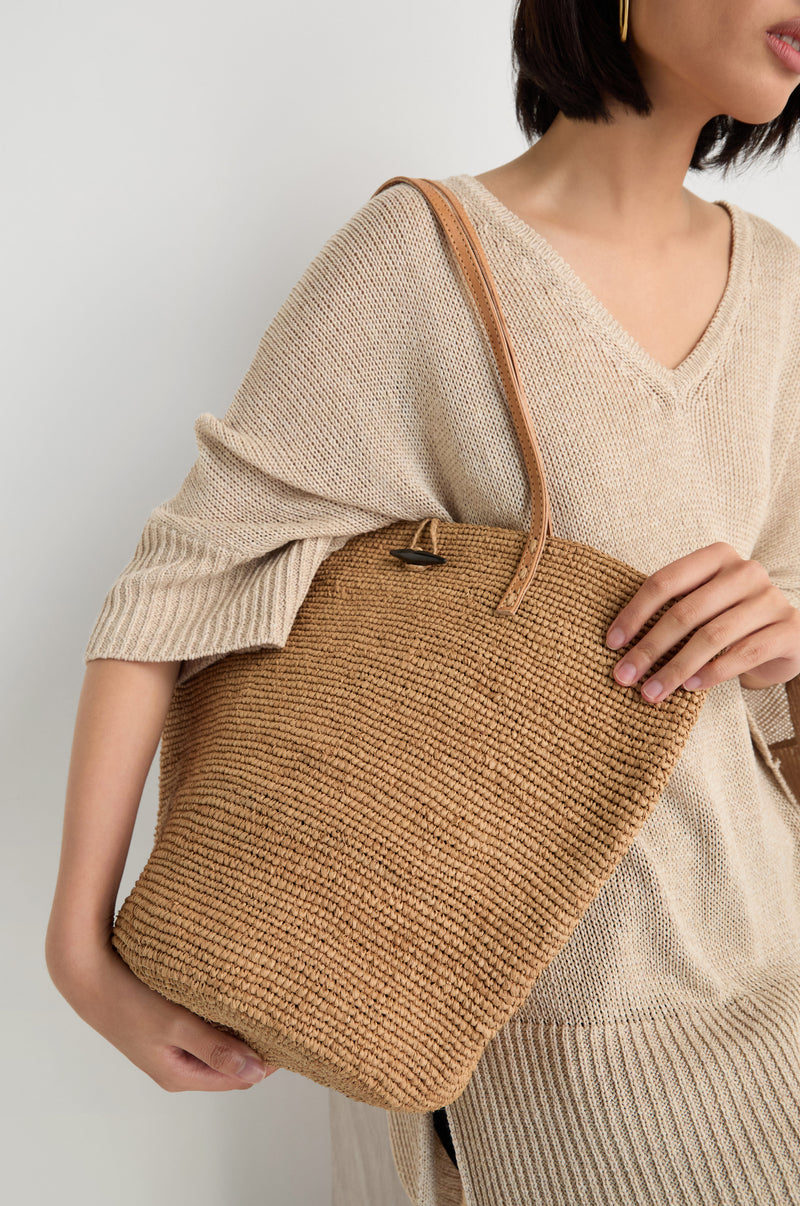 A woman in a beige knit top carries the Ilana Raffia Basket with leather handles over her shoulder against a plain background.