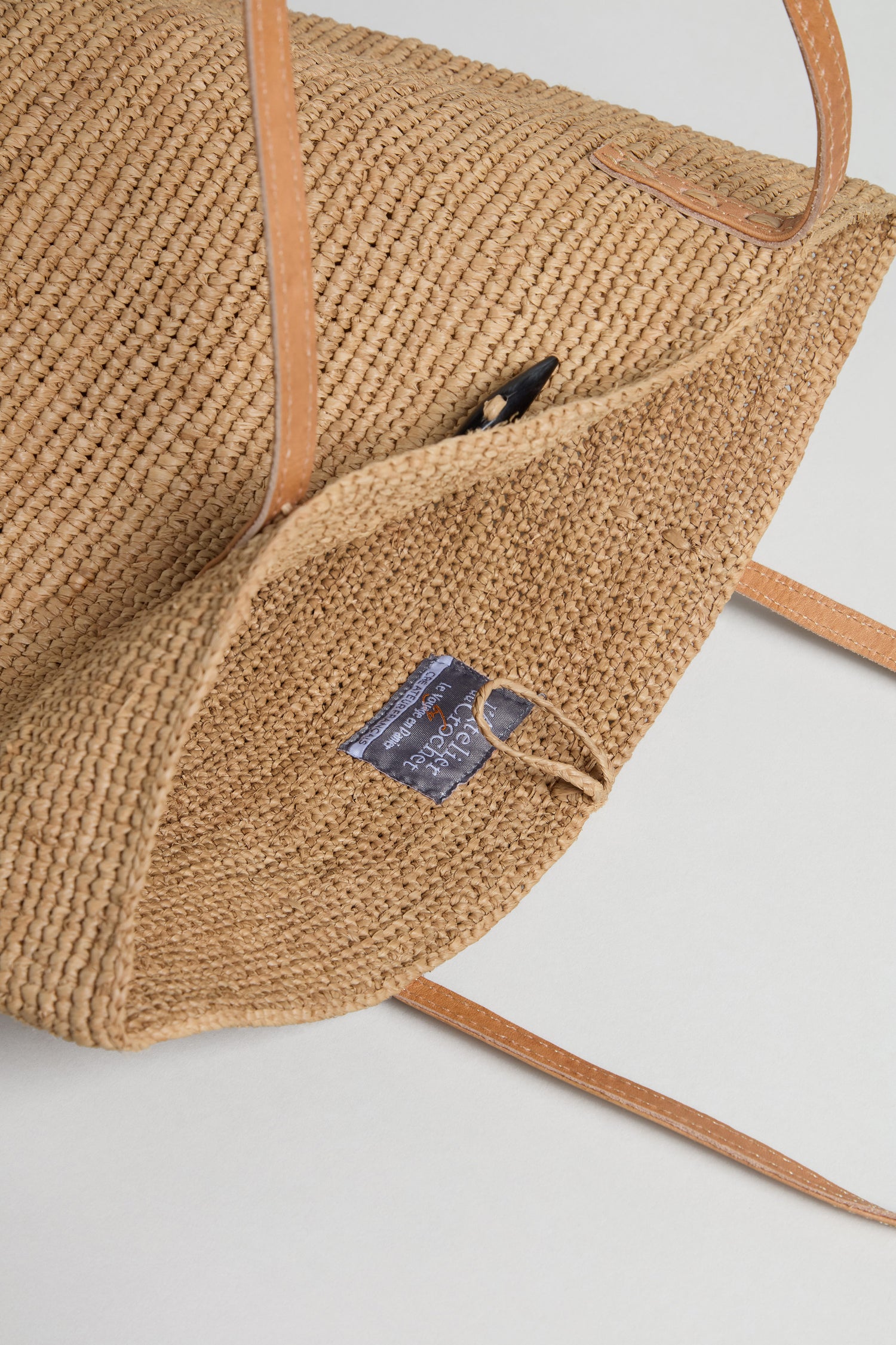 A close-up of the Ilana Raffia Basket, a woven straw tote with tan leather handles, interior label, and button closure loop—perfect for everyday use.