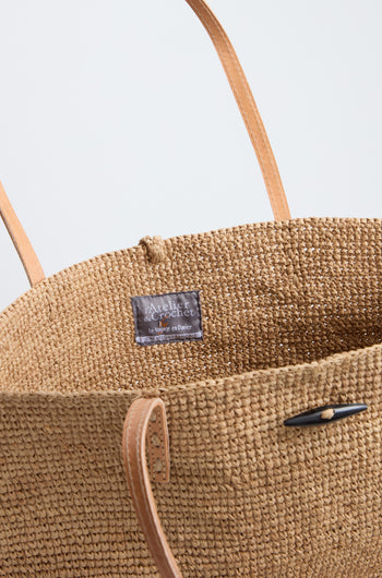 A close-up of the Ilana Raffia Basket, featuring handwoven design, tan leather handles, and a visible gray fabric label reading "Atelier du Crochet Le Voyage en Panier.