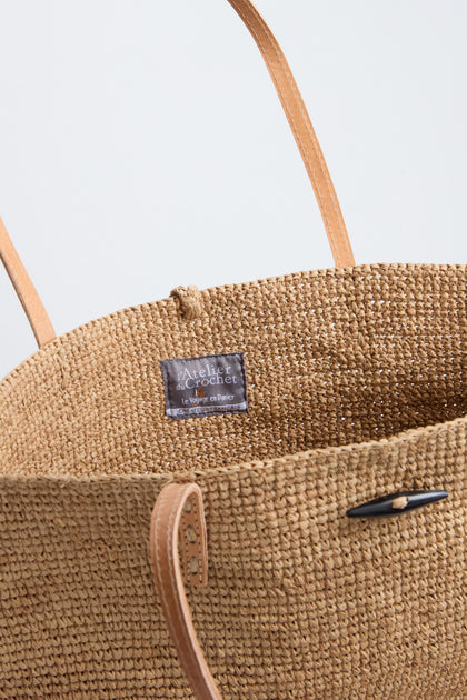 A close-up of the Ilana Raffia Basket, featuring handwoven design, tan leather handles, and a visible gray fabric label reading "Atelier du Crochet Le Voyage en Panier.