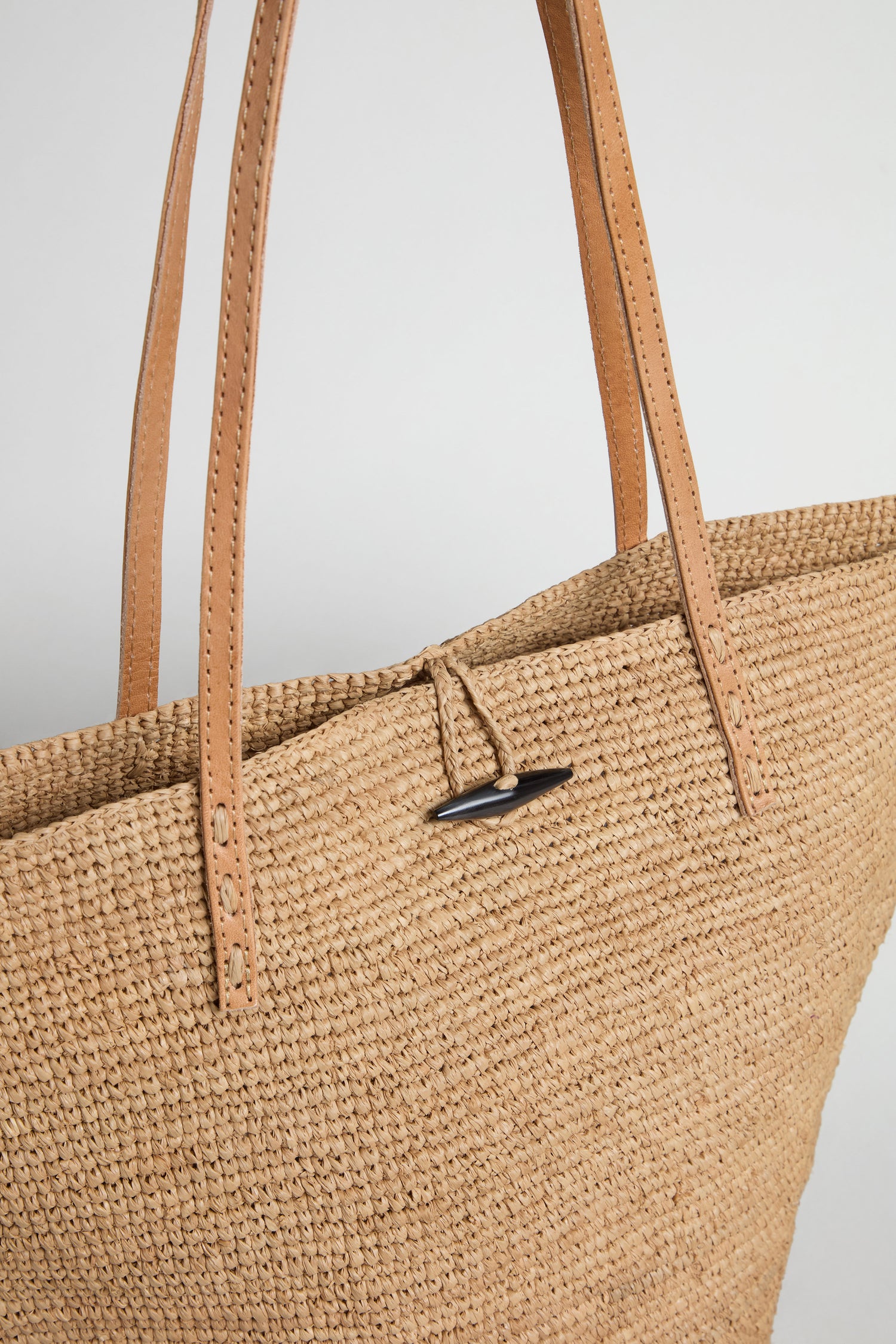 Close-up of the Ilana Raffia Basket, an everyday tote with a woven straw body, tan leather handles, and a black toggle closure.