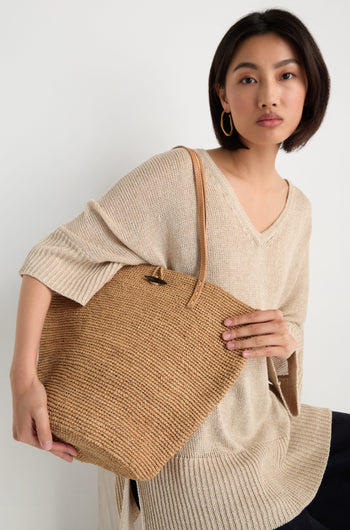 A woman in a beige sweater and gold hoop earrings holds the Ilana Raffia Basket, a large handwoven tote with leather handles, while seated against a plain background.