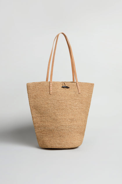 The Ilana Raffia Basket is a woven tote bag with two tan leather handles, shown upright on a plain white background—perfect for adding effortless style to your everyday look.