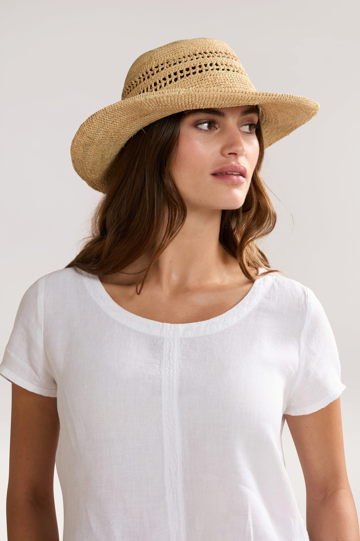 A woman in a white short-sleeved top and a Chapeau Westoloa, essential pieces of any summer wardrobe, looks to the side against a plain background.