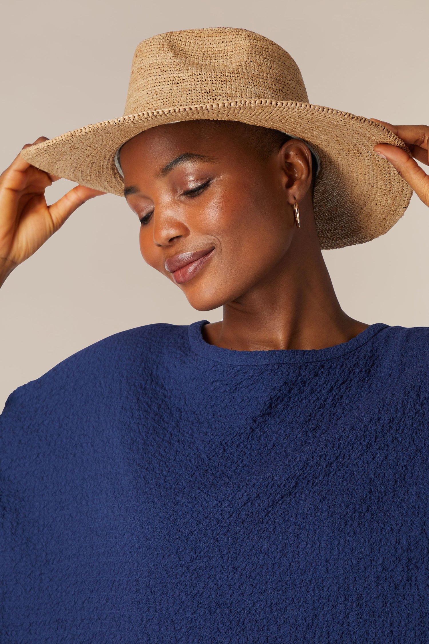 A person wearing a blue top and a straw hat smiles with eyes closed, holding the brim of their Chapeau Westilux with both hands—an ideal snapshot of a perfect summer wardrobe.